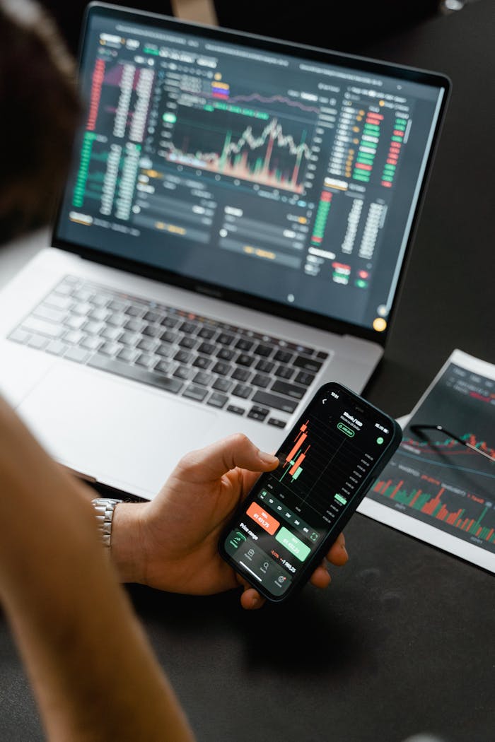 Suporte e atendimento da FxInnova: como falar com a equipe e resolver dúvidas Person analyzing stock market data on a smartphone with a laptop displaying financial graphs in the background.